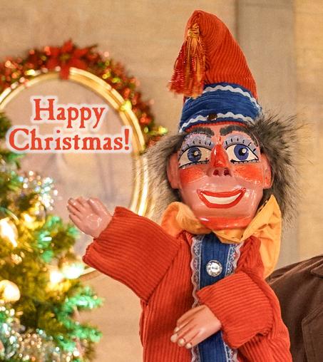 A festive looking, carved wooden Mr. Punch puppet with a wide-eyed, smiling face smiles and waves.  The words "Happy Christmas" are along side him. In the background there is a lighted, decorated Christmas tree.