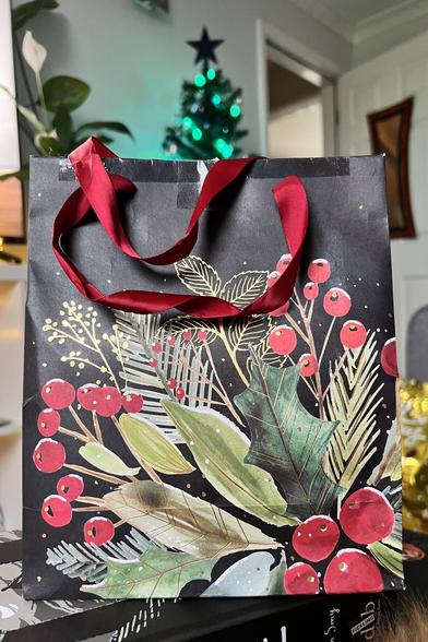 A festive gift bag, black in colour with green and red Christmassy foliage depicted.