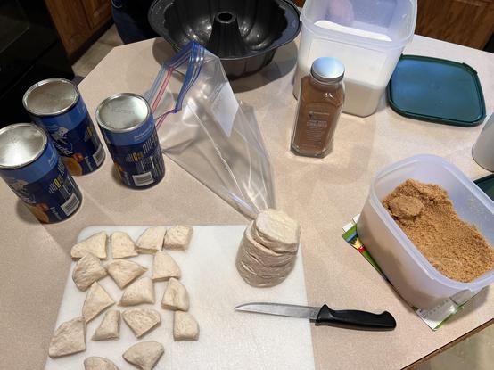A photo of a center kitchen island, with a lighter colored counter color than you usually see in my photos, with a bunch of different parts of a recipe gathered on it. Visible are tubes of buttermilk biscuits, a jar of cinnamon powder, and open containers of white sugar and brown sugar. There is a cutting board with some biscuits on it cut into fourths, an empty gallon ziploc bag, and a Bundt pan in the background.
