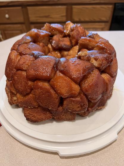 A photo of freshly baked monkey bread - a Bundt cake shaped dough, with bits of biscuit baked and held together by a brown sugar mixture. The monkey bread is setting on a cake carrier plastic base.