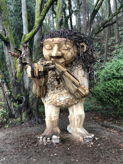 A wooden sculpture of a giant troll with a peaceful aspect. The troll is playing a huge flute made out of a driftwood log.