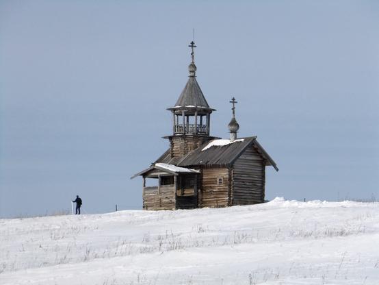A wooden chapel with a bell-tower on an empty snow-covered ground. A silhouette of a cross-country skier at the left of the chapel.
