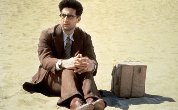 Man in a suit sitting on the sand with a wrapped box beside him.
