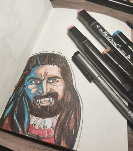 Foto of a sketchbook. Shows a finished illustration in European comic style. A m with dark brown long hair and a full beard. He has fangs. The person being Nandor from the show What we do in the shadows