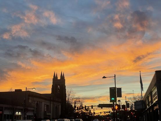 A band of clouds going from left to right, colored orange/red/even a little pink? from the setting sun, which is not visible.  Other scattered clouds as well against a blue sky.  Silhouetted by the clouds is a gothic-style church steeple with 4 small spires at the top, other buildings, street lights, traffic lights, cars...

(the only really visible lit traffic signal is a green bike lane signal. Yay bikes!)
