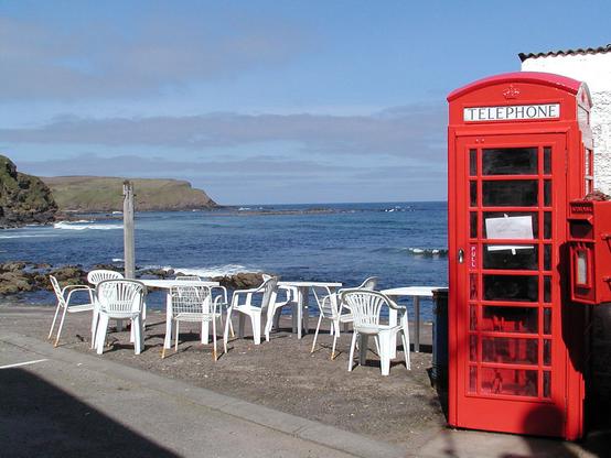 The image shows a red telephone box and post box on the right of the frame, with some white plastic tables and chairs on a quayside across the lower quarter of the frame. In the background is a blue sea with breaking waves and rockyheadlands on the left. The sky is mainly blue.