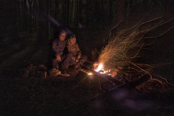 Dvě postavy, asi táta se synem, sedí v noci u ohně a opékají buřty. Všude okolo temný les, který ozařuje světlo ohně.