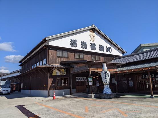 Yuasa Soy sauce 2 storey building with dark wooden siding and white top that has Japanese letters with the name. A tall (8 feet) white porcelain bottle of soy sauce is outside the entrance