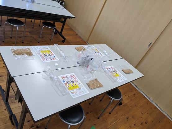 A table and stools in room. The table is set up with 4 stations with empty bottles and bags of soy sauce dry ingredients and instruction sheet