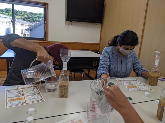 Two kids sitting across a table, reading instructions and pouring and measuring water and with soy sauce ingredients in bottles in front of them