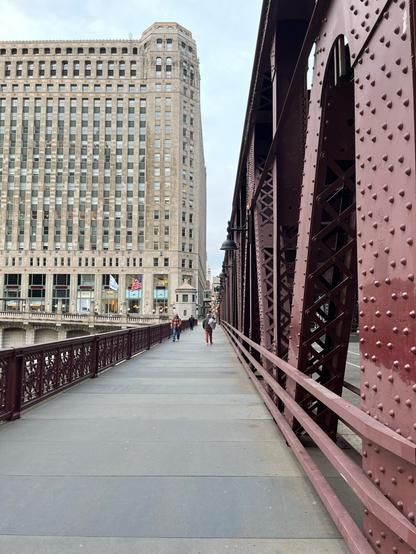 A photo of a walkway over a river. On the right side is a reddish brown railway bridge with A frames. At the far end is a very large Art Deco building at least 20 stories tall.