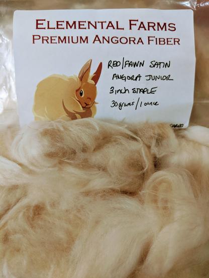 A pile of reddish Angora wool, next to a label that reads "Elemental Farms, premium Angora fiber. Red / fawn satin Angora junior. Three inch staple, 30 grams / 1 ounce." there's a cute drawing of a rabbit on the label
