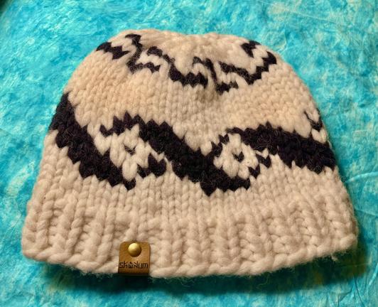 An off-white soft wool hat with design in black rests on a turquoise green cloth.  The hat has a tiny soft leather label identifying the designer, "Skukum Design."