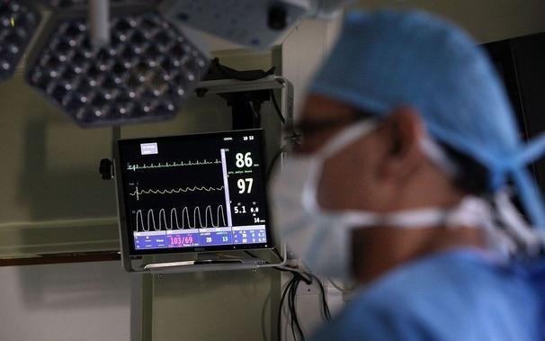 photo of a white, male medical professional dressed in blue scrubs and a white face mask, standing in front of a display screen with blood pressure, pulse, etc. readings