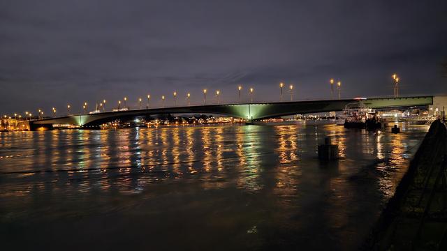 A photo of the Rhine at night, the colorful light from the buildings opposite is reflected in the surface. a large bridge leads to the other side and is also reflected