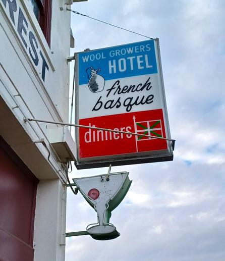 Red, white, and blue vintage sign attached to the side of an old, stucco-coated building: Wool Growers Hotel, French Basque, Dinners (with a red, green, and white Basque flag). A smaller sign in the shape of a martini glass outlined in neon is below.