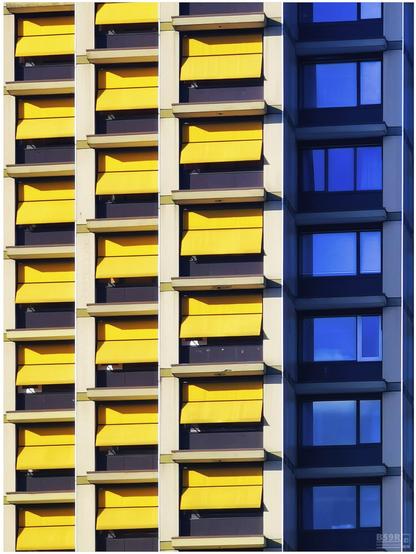 Das Foto zeigt im Hochformat über sieben Stockwerke die Balkonfassade von 3 nebeneinander stehenden und jeweils leicht nach hinten versetzten Wohntürmen.  Die Balkone sind zur Abschattung mit knallgelben geöffneten Markisen versehen. Am rechten Bildrand schliesst sich - ebenfalls etwas nach hinten versetzt - ein weiterer Wohnturm mit Balkonen an, jedoch fehlen hier die gelben Markisen, so dass sich der blaue Himmel in den großformatigen Fensterflächen spiegelt, wodurch sich ein reizvoller Farbkontrast zu den gelben Markisen der nebenliegenden Gebäude ergibt.

The photo shows the balcony façade of 3 neighbouring residential towers, each slightly offset to the rear, in portrait format over seven storeys.  The balconies are shaded by bright yellow open awnings. Another residential tower with balconies is located on the right-hand edge of the picture - also slightly offset to the rear - but here the yellow awnings are missing, so that the blue sky is reflected in the large windows, creating an attractive colour contrast to the yellow awnings of the adjacent buildings.