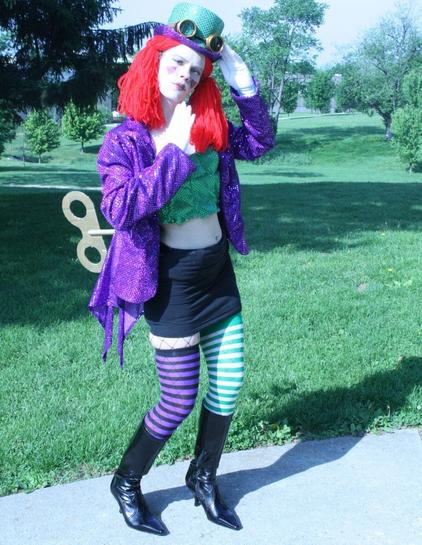 An actor poses for a publicity photograph outdoors near a park dressed as a parody of Columbia from "The Rocky Horror Picture Show" in green and purple sequins as a steampunk clockwork maid with goggles, a giant wind-up key, and 19th pantomime make-up.