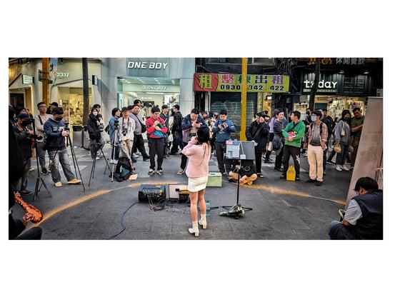 A public street. A woman is standing with her back to the viewer in the foreground. She is singing. There are several male spectators filming her.