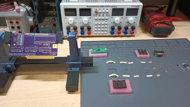 Bare PCB in a board holder, sitting on a workbench.  Laid out beside are resistors, capacitors, sockets, chips, and a Raspberry Pi Pico, waiting to be installed on the PCB.