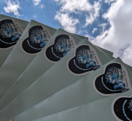 A photo of the corners of several fanned out envelopes against the backdrop of a cloudy blue sky. The envelopes bear custom heart-shaped postage stamps of a blue tiger.