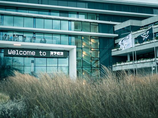 Foto vom Eingangsbereich des CCH (Congress Center Hamburg). Im unteren Drittel des Bildes leicht verschwommenes trockenes Gras. Im Fokus die spiegelnde Fensterfläche des Kongress-Gebäudes mit dem "Welcome to 37C3" Schild und den schwarzen und weißen Flaggen des Chaos Computer Club.
