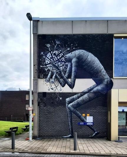 Streetartwall. A black and white mural of a large dark shadow figure was sprayed/painted on the exterior wall of a multi-story modern cultural building. The figure walks hunched over, clutching its head with its hands, which consists of antennae, planets on tree trunks, galaxies, stars and circles. Her eyes are two old, long binoculars.
Info: The anonymous Phlegm is a special street artist because he has developed his very own style based on comic figures he has already drawn. His apocalyptic figures become part of the architecture on which they live. Phlegm comes from Nordwaldes. The name Phlegm comes from one of the four humors in ancient Greek medicine: blood, yellow bile, black bile and phlegm. It was believed that phlegm was responsible for an apathetic and emotionless temperament.