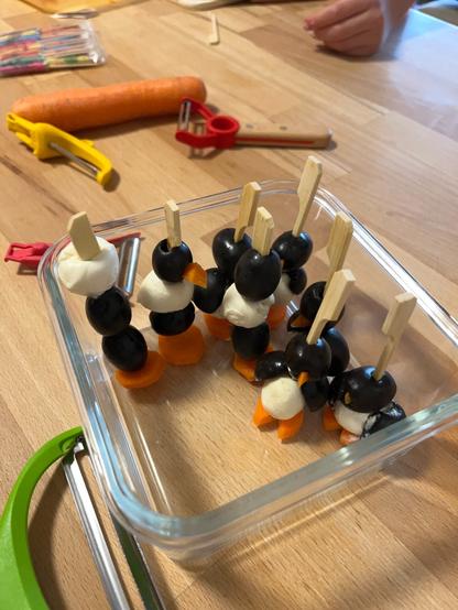 Ein paar „Pinguine“ aus Mozzarellakugeln, Karottenbeinchen- und schnäbeln und Olivenfrack und -Kopf mit Holzspieß in einer viereckigen Glasschale.