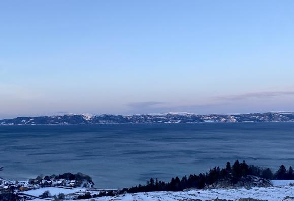 View of the Trondheimsfjord and the land on the other side (Fosen) in low sunlight. Blue colors with a hint of pink above the low mountains.