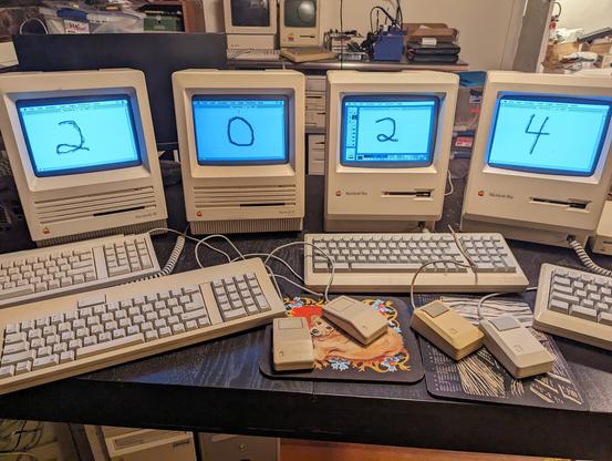 Four compact Macs (two SE and two Plus) lined up on a table, with keyboards and mice in front. The screens spell out "2024" via a MacPaint document in each.