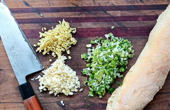 Vorbereitung zu dem Gericht:
Ein Holzbrett mit einem großen japanischen Messer, ein selbstgebackenes Baguette und größere Menschen klein geschnittene Frühlingszwiebeln, Knoblauch und Ingwer