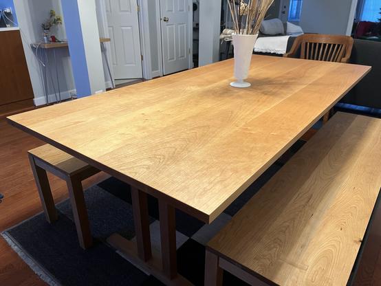 Wooden dining table with a white vase, set in a room with surrounding furniture and doors in the background.