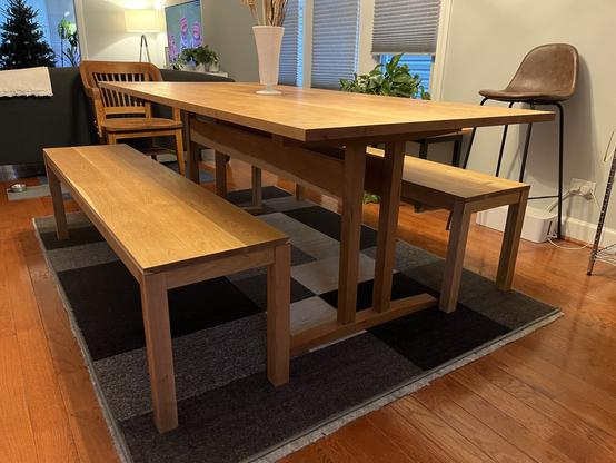A dining room with a wooden table and benches on a grey and black area rug, a wooden chair, a brown leather chair, and a plant on the window sill.