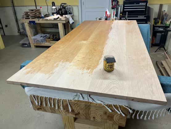 Wooden tabletop undergoing finishing with a jar of Odie’s oil on it, placed on a workbench with woodworking tools and work items in the background.