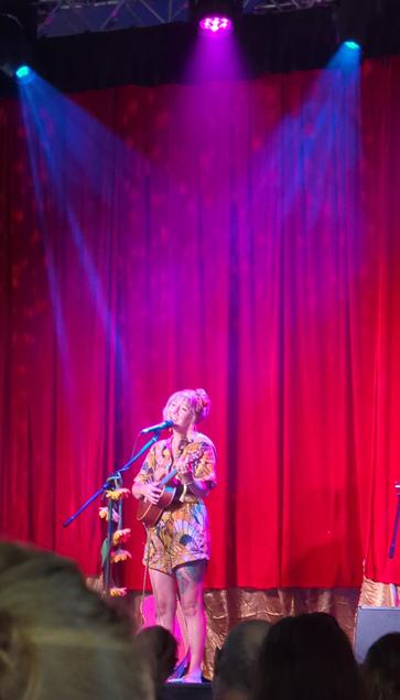 Alana Wilkinson on stage at the Parlour, standing in front of a dark red curtain in matching bright yellow shorts and shirt, singing into a microphone while playing ukelele.