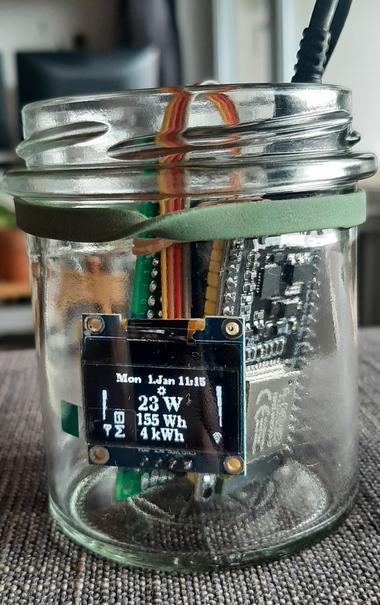 Das Bild zeigt ein paar bestueckte Platinen inkl. einem kleinen Display, das mit einem Gummiband zusammehalten wird und in ein offenes Twist-off Glas gestellt wurde. AUf den DIsplay steht:
Mon 1. Jan 11:15
23W 
155 Wh 
4 kWh