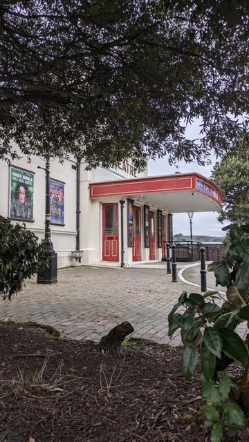 The front entrance of a theatre with red doors and red overhead panel. Brick paving leading to the entrance with posters displaying future events on the wall to the left