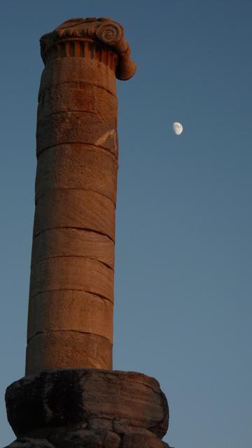 An unfinished Ionic column, tinted orange by the setting sun, rears up into the evening sky, in which hangs a gibbous moon.