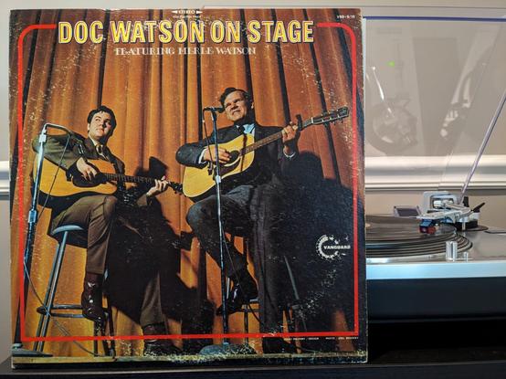 LP spinning on turntable: DOC WATSON ON STAGE FEATURING MERLE WATSON. Cover w/ photo of Doc & Merle playing guitars on stage.