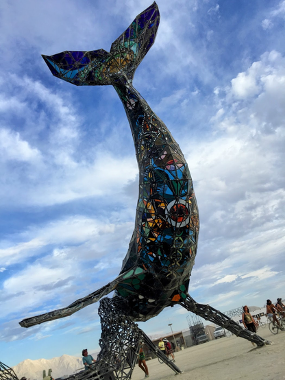 a photograph of a humpback whale made of stained glass silhouetted against a blue and lightly cloudy sky. At the base of the sculpture, a few people gather; they are smaller than the whale's fin.