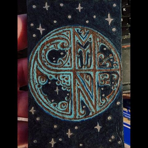 A photo of a trading-card-sized coloured pencil drawing held in the artist's hand.  The drawing is of a round sigil in copper filigree with a verdigris patina against a starry sky.