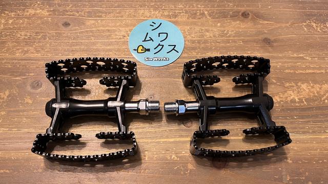 A pair of black bicycle pedals on a wooden surface accompanied by a round blue sticker with Japanese text and a cartoon character.