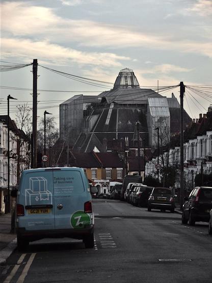 View down a late Victorian street of terraced houses. Closest, a light blue hire van is parked, facing away, on the left. Telephone cables stretch across between wooden poles on either side, and radiate out to each house. At the far end of the street, beyond the houses, is an immense dark grey roof, made from many different planes, three times the height of a the houses, and eight times as wide. Two glass towers (lift shafts?) rise from the edges of the roof. A faceted glass cupola tops the construction.