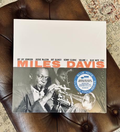 Photo of a record album jacket laying on a tan leather stool. 

The jacket is white and the lower half a monochrome photo of Miles Davis blowing into his trumpet. 

Orange text runs across the top of the photo and reads, ‘Miles Davis’. The line up is listed in black type directly above. 

A blue and white hype sticker reads, ‘Blue Note Classic Vinyl Series’ to the right of the photo.