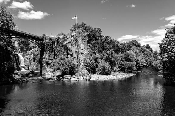 A black and white photograph showing a waterfall on a sunny summer day. Taken at a bend in the river at the bottom of the falls, a tall cliff face forming the inside of the bend is in the center, an American flag raised on the promontory. To the left, upriver, a waterfall is visible, water descending from the same level as the cliff downward. A metal footbridge crosses the river between the promontory and the cliff wall on the opposite (left) side. On the right side of the bend, water filling the foreground, the flow continues downriver, lined with trees.