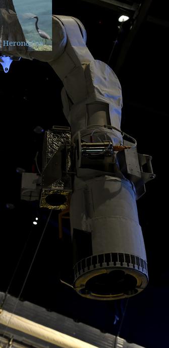 An indoor photograph of the end of a robot arm. The end is a hollow cylinder with a TV camera near the end, and two joints covered in a white, fabric-like material are above the end effector. Abover it are circular panels joined by rectangular sections all coveted in white cloth.