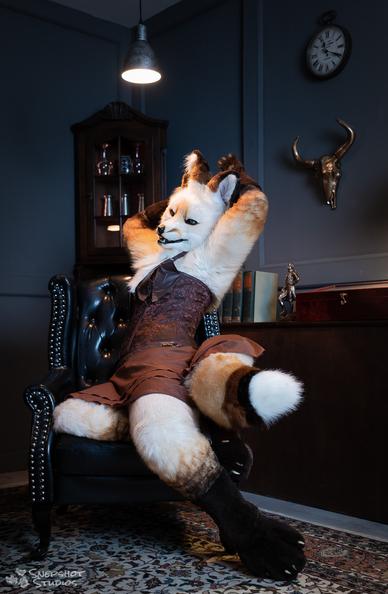 White vixen fursuit in a dark dress lounging in an old leather chair.