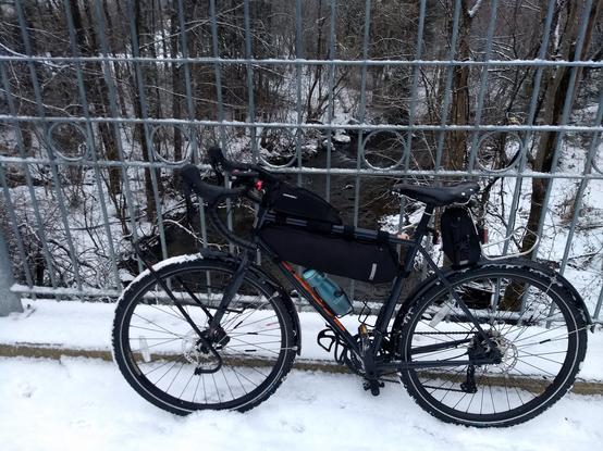 [fr]Un vélo gravel gris foncé à pneus d'hiver appuyé sur la clôture grillagée d'un petit pont. Une rivière bordée d'arbres est visible en arrière-plan, la neige recouvre le sol. [en]A dark gray gravel bike leans on a small bridge's steel fence. A river bored by trees is seen in the background, snow covers the ground.