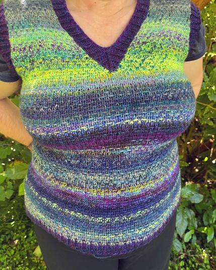 Torso shot of a hand knitted vest with a woven look slip stitch motif. The vest includes yarn shades from chartreuse to blue to purple and has bands of dark purple.