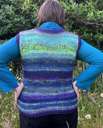 The back of a person wearing a hand knitted vest with a woven look slip stitch motif. The vest includes yarn shades from chartreuse to blue to purple and has bands of dark purple. Behind the person is grass and trees.